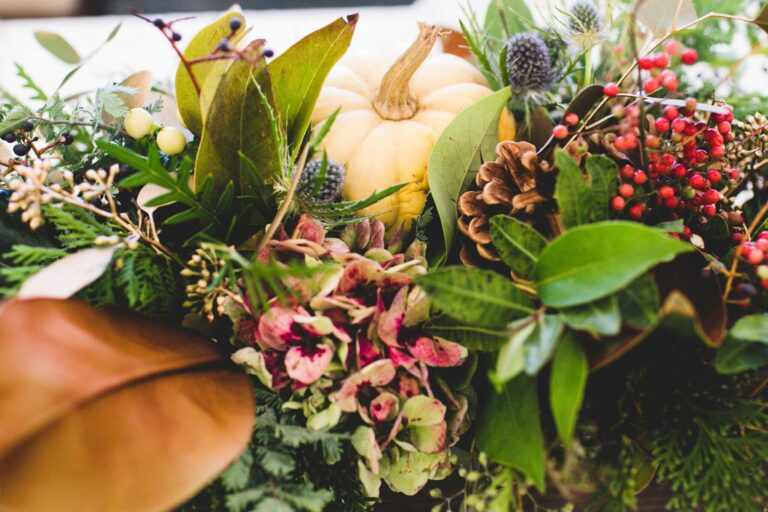 A vibrant arrangement of autumn foliage, featuring a small pumpkin, pinecones, and colorful berries among lush greenery.