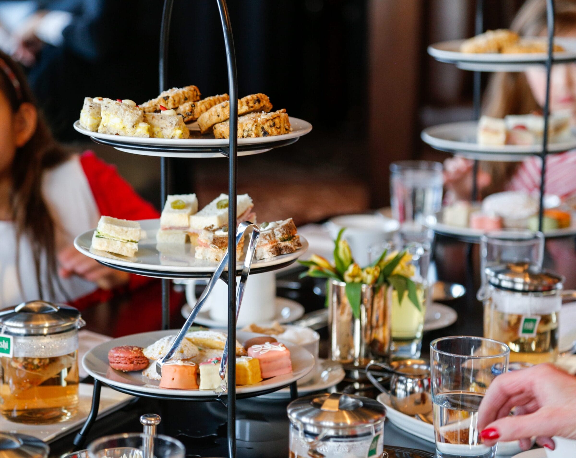 Three-tiered stand filled with assorted sandwiches and pastries, accompanied by tea and drinks on a dining table.