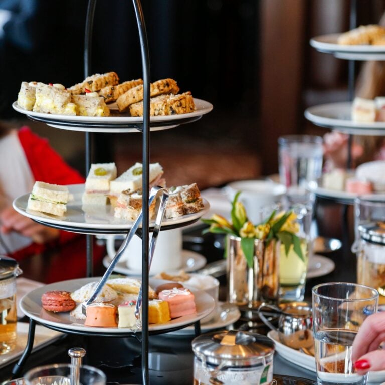 Three-tiered stand filled with assorted sandwiches and pastries, accompanied by tea and drinks on a dining table.